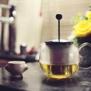 A glass teapot of green tea beside a small cup and yellow roses