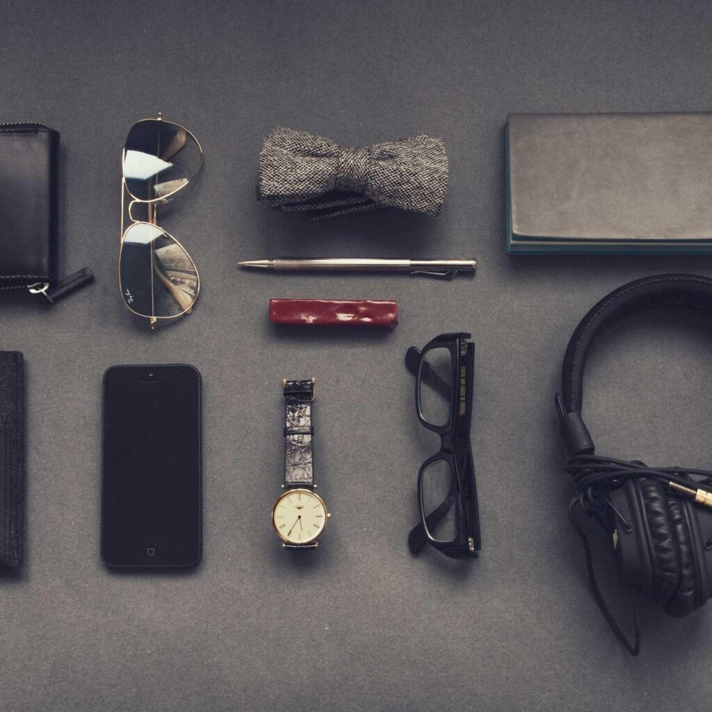 Travel essentials laid out on grey: wallet, sunglasses, phone, watch, glasses and headphones